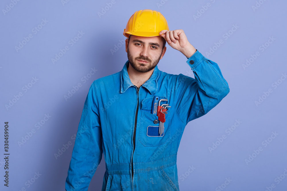 Portrait of serious responsible young worker holding his yellow helmet with one hand, wearing uniform, having equipment in pockets, standing isolated over blue background in studio. Work concept. Portrait of serious responsible young worker holding his yellow helmet with one hand, wearing uniform, having equipment in pockets, standing isolated over blue background in studio. Work concept.
