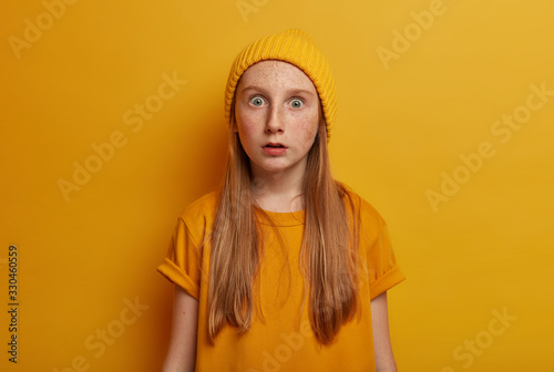 Startled amazed small girl has freckled face, stares with bugged eyes, sees something impressed, wears yellow hat and t shirt, cannot believe in shocking relevation, realizes terrible situation © Wayhome Studio
