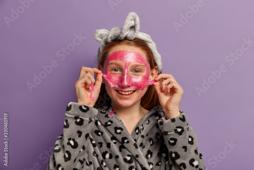 Indoor shot of happy female child takes off facial mask, cares about complexion, has postive smile, wears casual robe, poses against purple background, prepares for first date with boyfriend