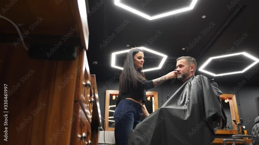 Barber woman cuts bearded man's hair with a clipper in barbershop. Men's hairstyling and hair cutting in salon. Grooming the hair with trimmer. Hairdresser doing haircut in retro hair salon. Barber woman cuts bearded man's hair with a clipper in barbershop. Men's hairstyling and hair cutting in salon. Grooming the hair with trimmer. Hairdresser doing haircut in retro hair salon.