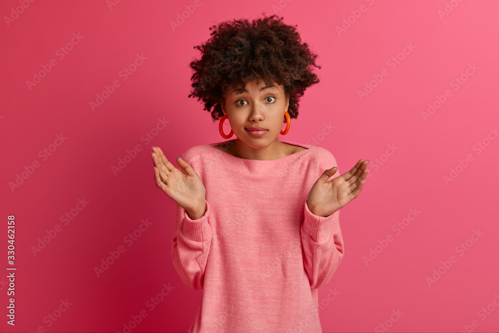 Indecisive curly young woman with dark skin shrugs shoulders, asks what did wrong, stands confused troubled and perplexed, keeps palms sideways, isolated over pink background, dressed casually