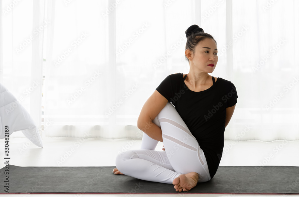 Fototapeta premium Asian women doing yoga seated twisting exercise at home, sitting in Lord of the Half Fishes Pose or Ardha Matsyendrasana, in white bedroom. Working out wearing black shirt and white pants