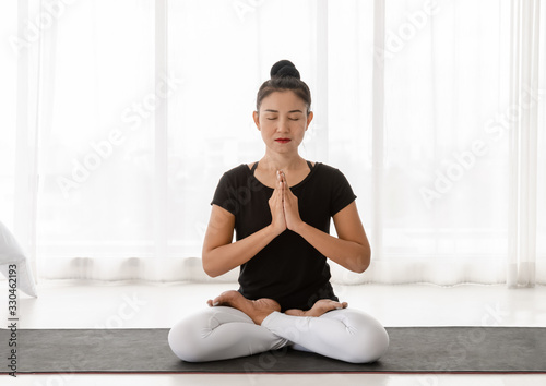 Asian women doing yoga meditating exercise at home, sitting in Lotus pose or Padmasana with raised hands namaste in white bedroom. Working out wearing black shirt and white pants. Healthcare concept..
