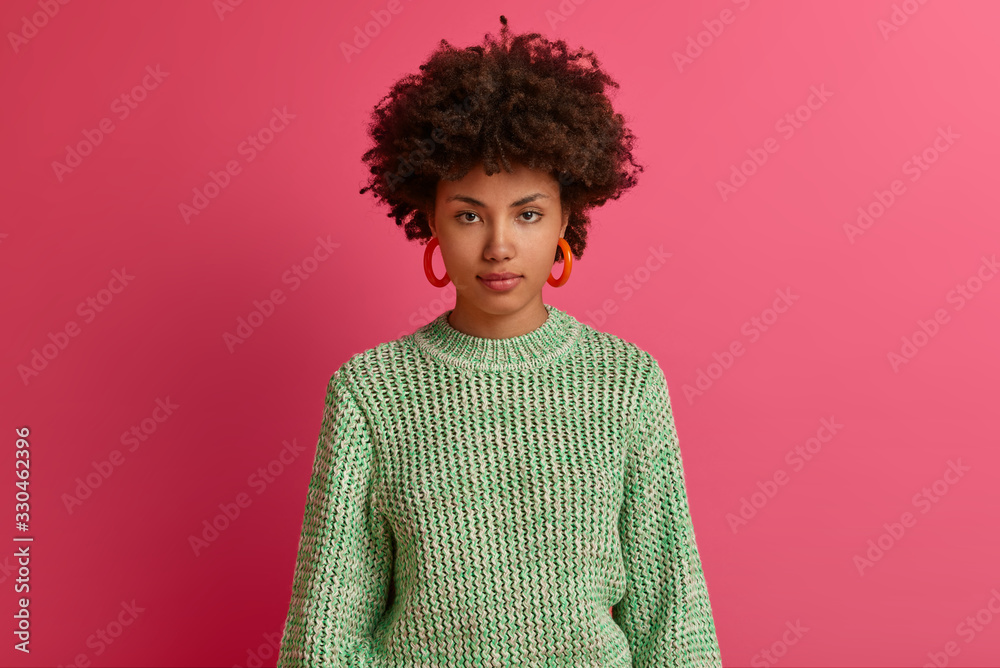 Confident serious determined young Afro American woman takes everything under control, listens information carefully, wears long sleeved knitted jumper, poses against pink studio background. Confident serious determined young Afro American woman takes everything under control, listens information carefully, wears long sleeved knitted jumper, poses against pink studio background.
