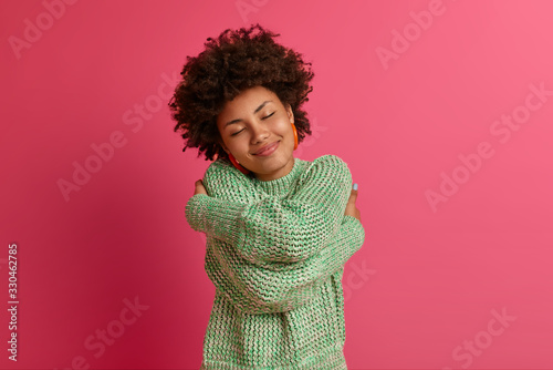 Konstfotografi Tender curly Afro American woman cuddles herself, closes eyes and tilts head, feels coziness, wears warm sweater, dreams about something pleasant, poses against rosy background