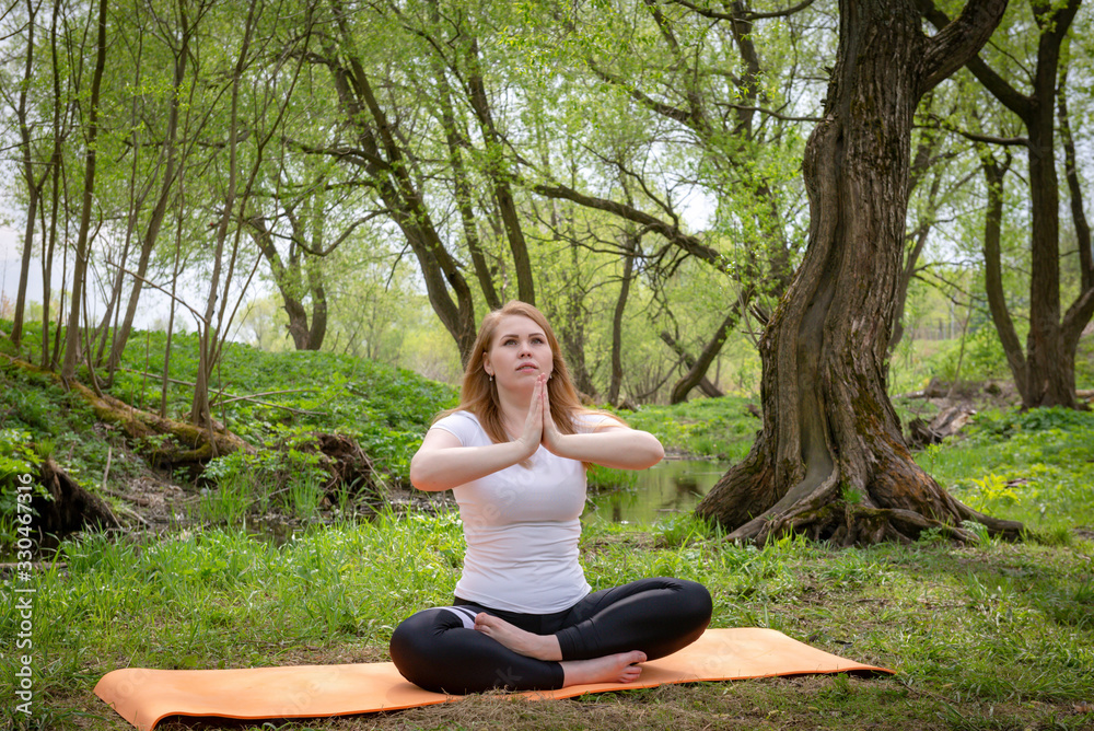 European young woman in a white top sitting on the grass in the Lotus position among the trees