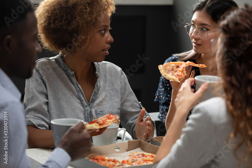 Diverse colleagues enjoying pizza together, African American and Asian businesswomen chatting during lunch break close up, employees team eating Italian fast food, talking, good relations