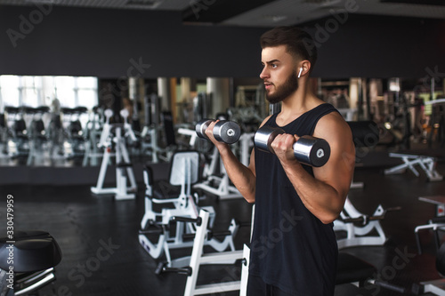 Muscular young man working out in gym doing exercises with dumbbells at biceps
