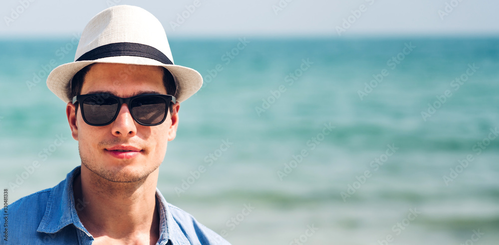 Portrait of smiling happy handsome man model enjoying and relax in fashionable sunglasses and hipster summer straw hat standing on the tropical beach and looking at camara.Summer vacations and travel