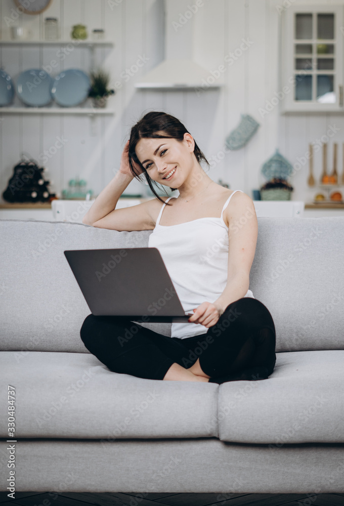 Fototapeta premium Excited brunette woman is sitting on the sofa in front of a laptop, reading media, smiling in living room