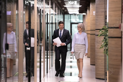 Smiling diverse businesspeople walking down modern hall or corridor in office, happy colleagues coworkers chat speak going to boardroom meeting or briefing together, cooperation concept