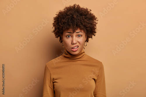 Dissatisfied dark skinned young woman purses lips, looks unpleasantly at camera, expresses aversion, smirks face, wears clothes, isolated on beige background. Negative face expressions concept © wayhome.studio 