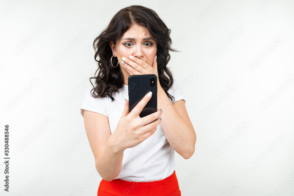 surprised girl covers her mouth with hand holding smartphone in front of herself on an isolated white background surprised girl covers her mouth with hand holding smartphone in front of herself on an isolated white background