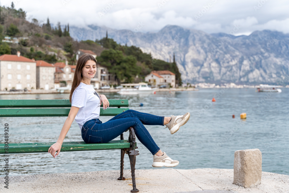 Fototapeta premium Woman in orange hoodie stands by the sea in city Perast on the background in Montenegro