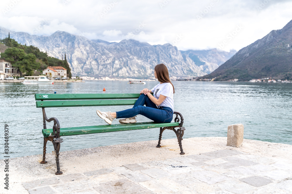 Fototapeta premium Woman in orange hoodie stands by the sea in city Perast on the background in Montenegro