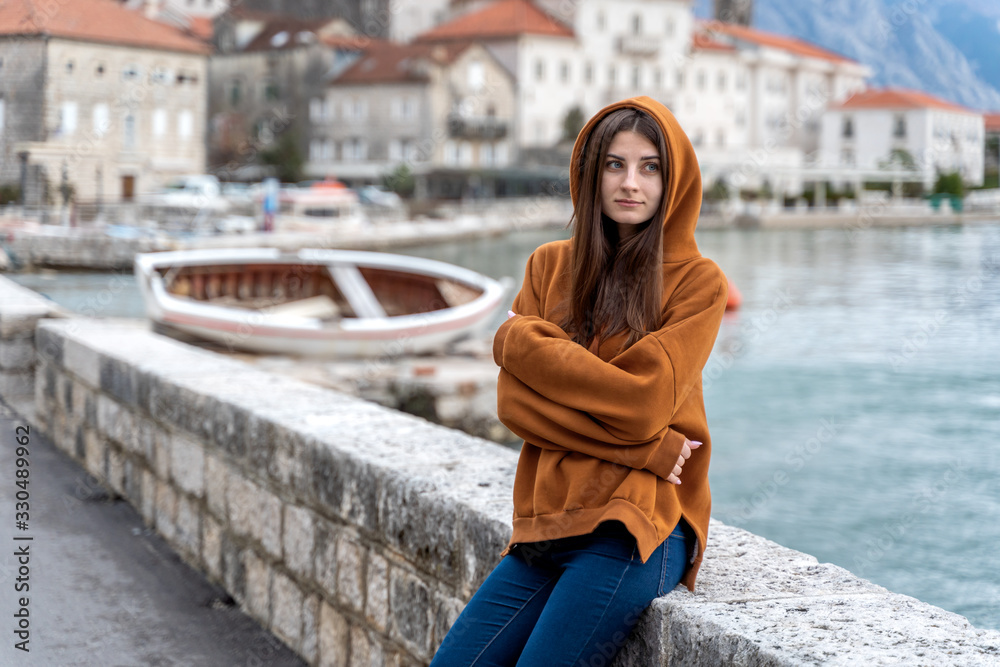 Fototapeta premium Woman in orange hoodie stands by the sea in city Perast on the background in Montenegro