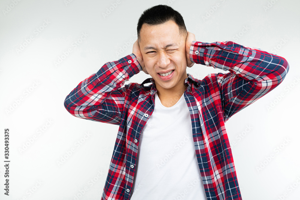 handsome asian guy in a checkered shirt wide open covers his ears from noise on a white studio background handsome asian guy in a checkered shirt wide open covers his ears from noise on a white studio background