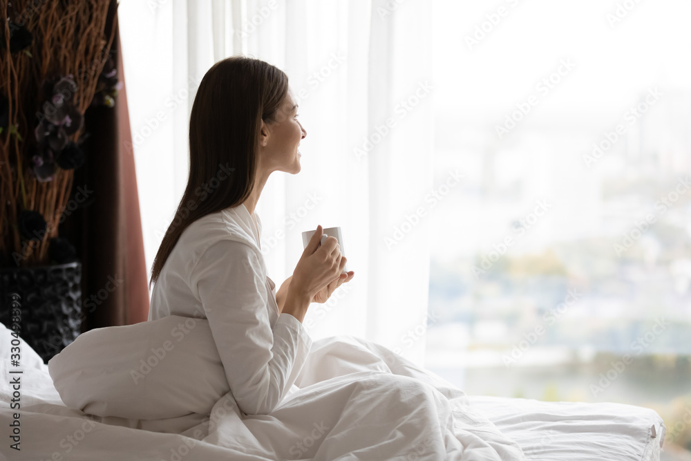 Side view happy attractive young brunette woman sitting in bed under duvet, holding cup of black coffee, enjoying peaceful calm weekend vacation morning time alone in bedroom at home or hotel. Side view happy attractive young brunette woman sitting in bed under duvet, holding cup of black coffee, enjoying peaceful calm weekend vacation morning time alone in bedroom at home or hotel.