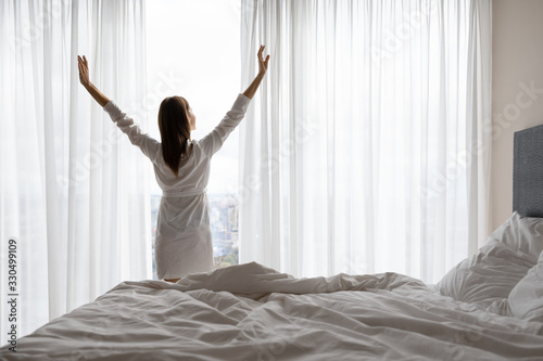 Rear back view young woman in night gown standing near panoramic window with opened curtains, stretching muscles after wake up. Energetic brunette lady doing exercises, enjoying good morning time.