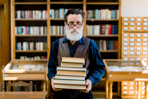 Foto Portrait of attractive bearded old man wearing shirt and leather vest, high school teacher or librarian, holding books in hands, standing in vintage library interior