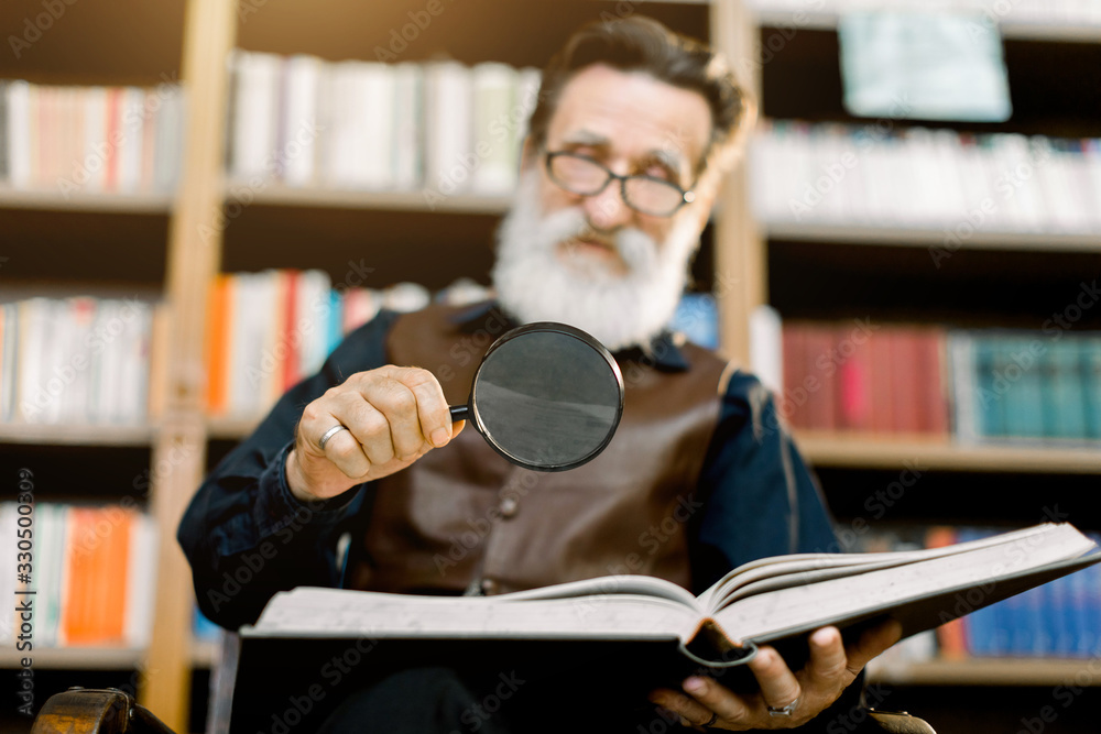 Φωτογραφία Handsome smiling senior bearded man, librarian or professor, in the library, sitting on the background of bookcases, holding magnifying glass and reading book