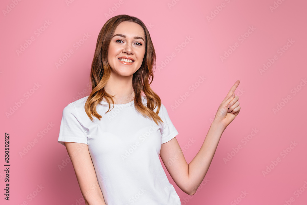 Happy student girl smiles and points a finger at empty copy space, close up. Portrait of a young woman isolated over pink background Happy student girl smiles and points a finger at empty copy space, close up. Portrait of a young woman isolated over pink background