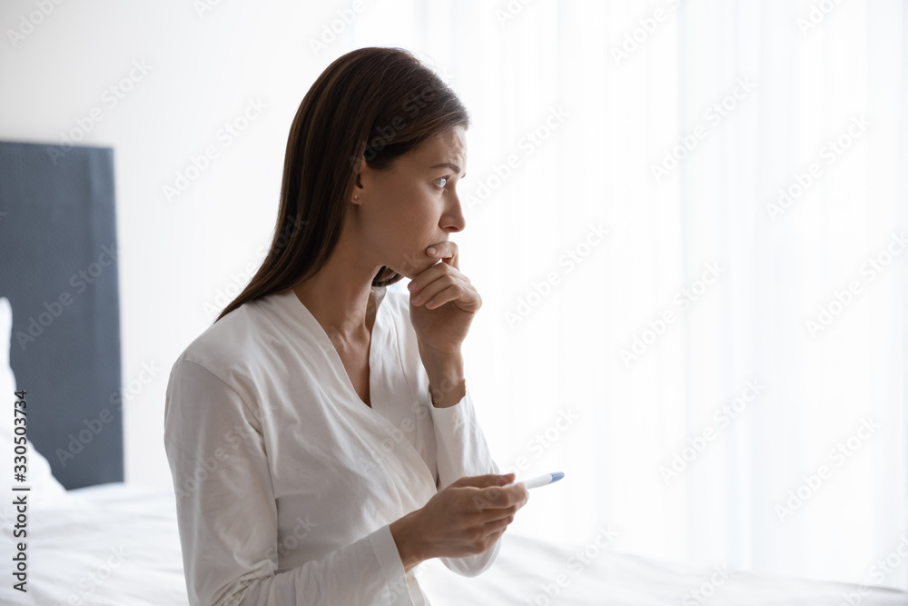 Head shot thoughtful young woman in nightgown bathrobe sitting on bed, holding quick plastic test in hands, worrying about infertility or unwanted unplanned pregnancy, future maternity concept. Head shot thoughtful young woman in nightgown bathrobe sitting on bed, holding quick plastic test in hands, worrying about infertility or unwanted unplanned pregnancy, future maternity concept.