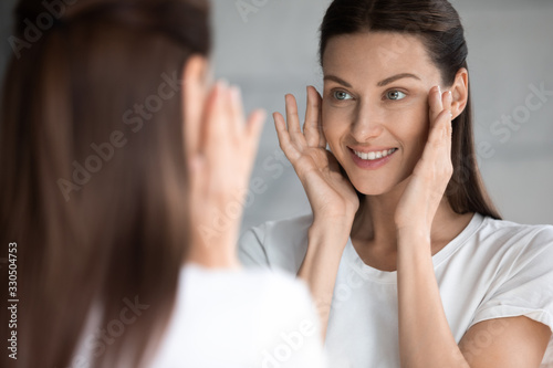Attractive smiling young brunette woman looking at mirror, touching face. Happy 30s lady satisfied with moisturized soft skin condition after professional cosmetology services, skincare concept. photo