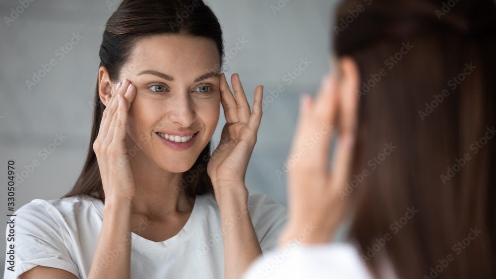 Close up head shot happy female client satisfied with eyes area condition after beauty spa procedure, looking in mirror at bathroom. Smiling pretty brunette woman moisturizing face skin in morning. Close up head shot happy female client satisfied with eyes area condition after beauty spa procedure, looking in mirror at bathroom. Smiling pretty brunette woman moisturizing face skin in morning.