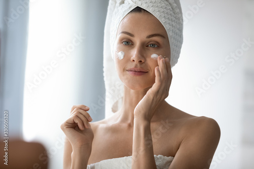 Head shot beautiful 30s lady looking in mirror, applying hydrating lotion creme on cheeks, finishing morning domestic skincare routine. Smiling woman grooming herself after showering in bathroom.