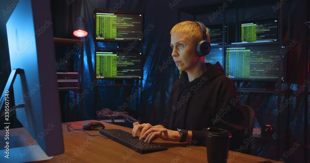 Caucasian woman hacker with blond short hair in headphones sitting in dark room with monitors at table and typing on keyboard of computer. Female hacking online system. Stealing information concept.
