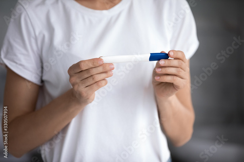 Cropped image young woman in white t-shirt holding quick pregnancy test, waiting for result. Focus on female hands with plastic stick, gynecologic healthcare, pregnancy confirmation or denial concept.