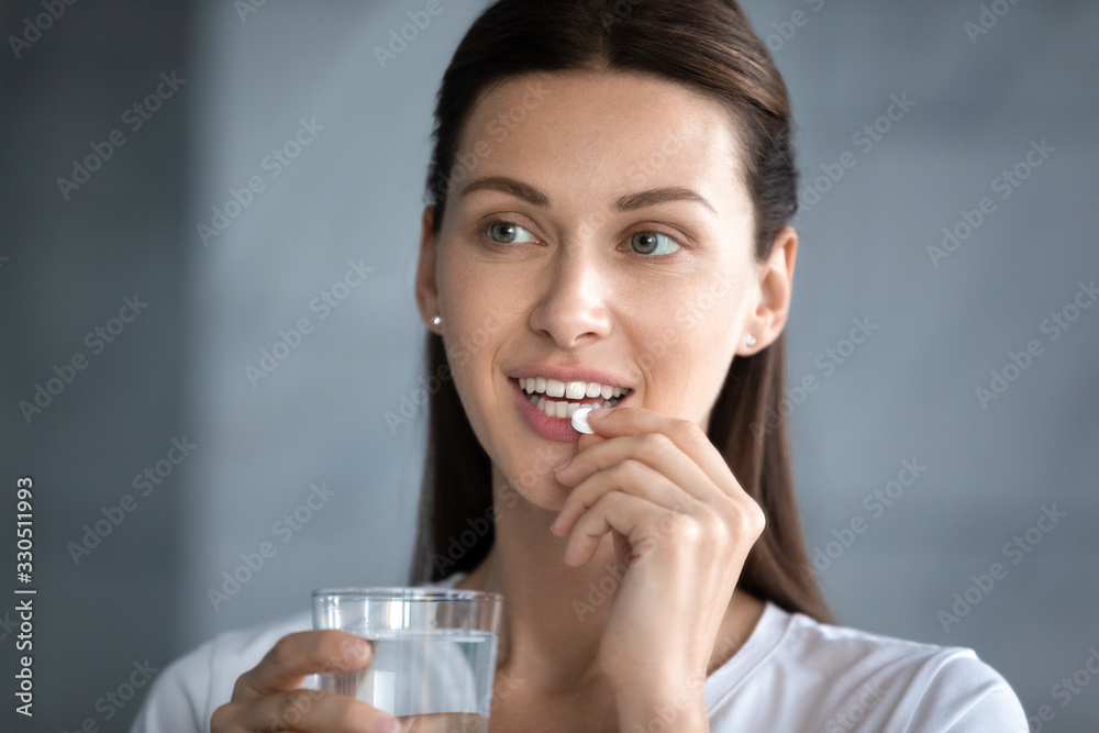 Close up head shot attractive brunette woman holding glass of mineral water, taking pill, looking away. Pretty young lady takes daily omega vitamins or dietary supplements for nails hair skincare. Close up head shot attractive brunette woman holding glass of mineral water, taking pill, looking away. Pretty young lady takes daily omega vitamins or dietary supplements for nails hair skincare.