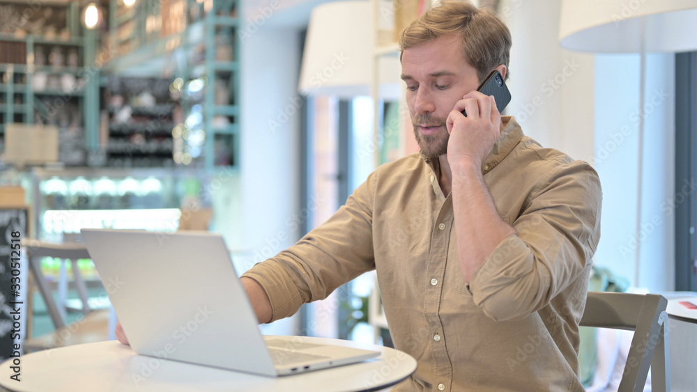 Young Man with Laptop Talking on Smartphone in Cafe