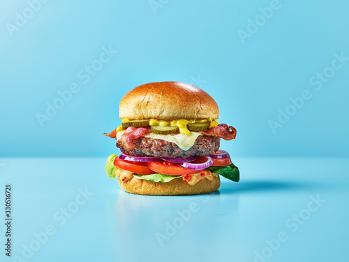 A cheese burger with bacon, salad, relish and gherkins against a blue background photo
