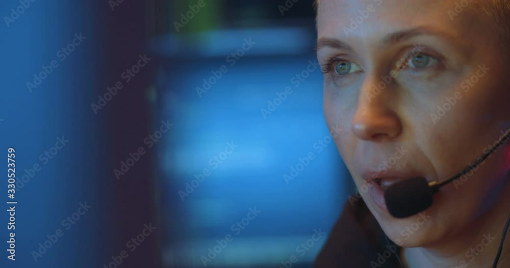 Close up of Caucasian middle-aged woman in headset with mic talking while having call with client in dark room with monitors. Female worker of call-center in front of computer screen.