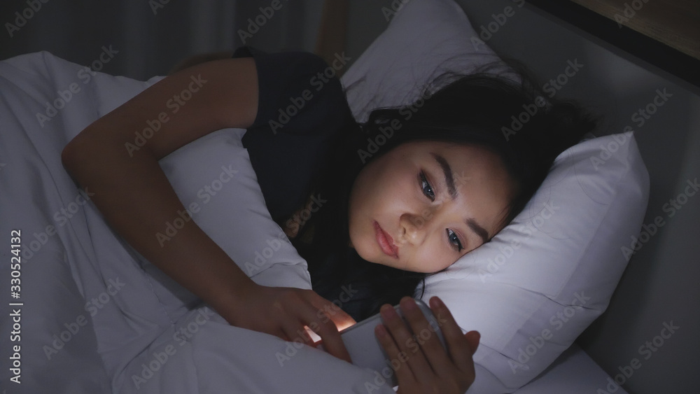 Asian young woman lying on bed play smartphones during night time In bedroom that turns off lights. Read COVID-19 on mobile phone. Using p hone in low light has health effect on eyes. Insomnia Concept - obrazy, fototapety, plakaty 