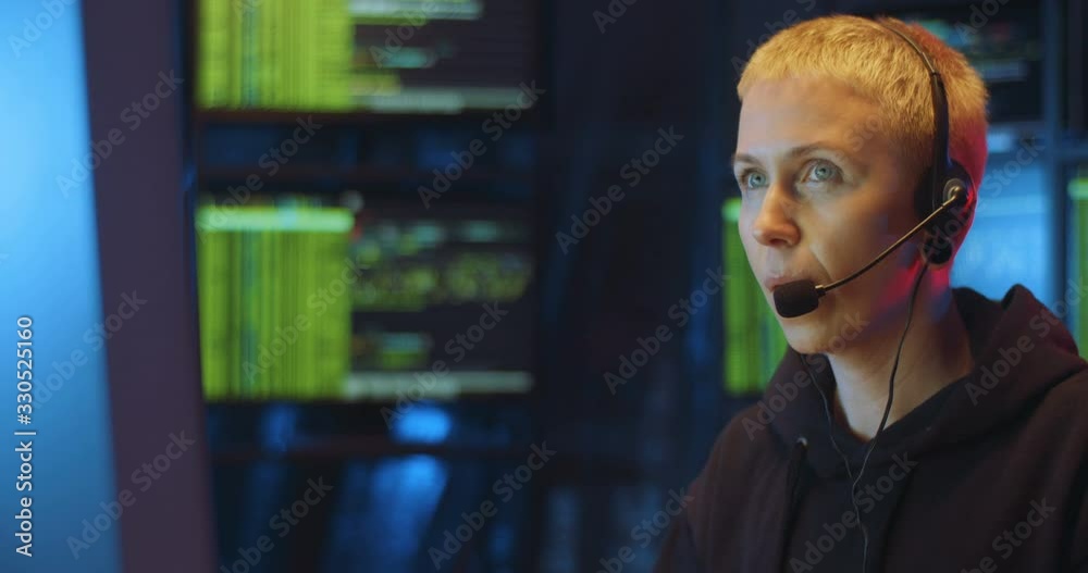 Caucasian woman with headset and mic sitting in dark room with monitors at desk and working at computer in call center at night. Female hacking online system. Stealing information concept.