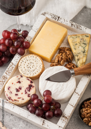 Glass of red wine with selection of various cheese in wooden box and grapes on light table background. Blue Stilton, Red Leicester and Brie Cheese with Cheddar and nuts with honey. Close up