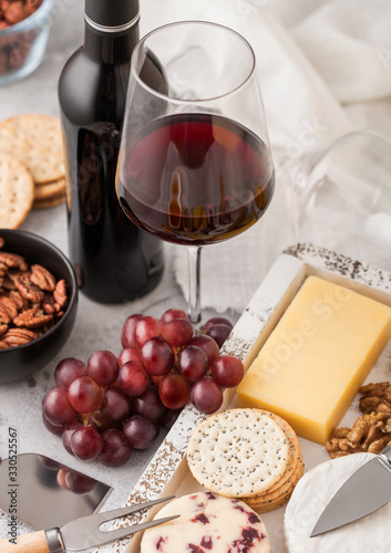 Bottle and glass of red wine with selection of various cheese in wooden box and grapes on light table background. Blue Stilton, Red Leicester and Brie Cheese with Cheddar and nuts with honey.