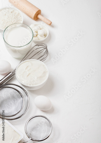 Fresh dairy products on white table background. Glass of milk, bowl of flour, sour cream and cottage cheese and eggs. Steel whisk and rolling pin. Top view
