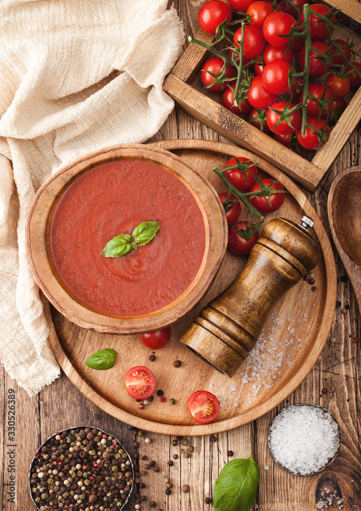 Wooden plate of creamy tomato soup 0n round tray, pepper and kitchen cloth on wooden background with box of raw tomatoes.
