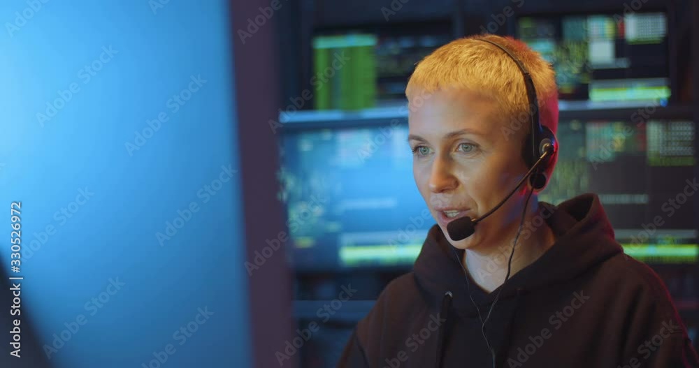Caucasian middle-aged woman with short blonde hair and in headset speaking in mic while working at compuer screen in call monitoring center in dark room at night. Female cyber security policewoman.
