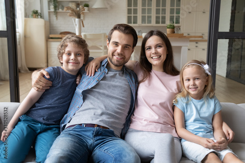 Portrait of happy parents hug cuddle with small kids relax on cozy sofa in living room, smiling young family with children sit rest on comfortable couch look at camera enjoying leisure weekend at home