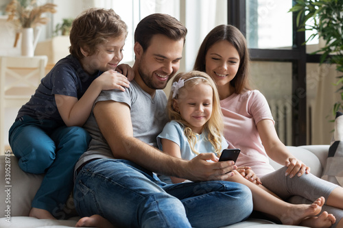 Happy young family with little children sit relax on comfortable couch watching funny video on smartphone together, smiling parents with kids rest on sofa at home enjoying cartoon on cellphone © fizkes