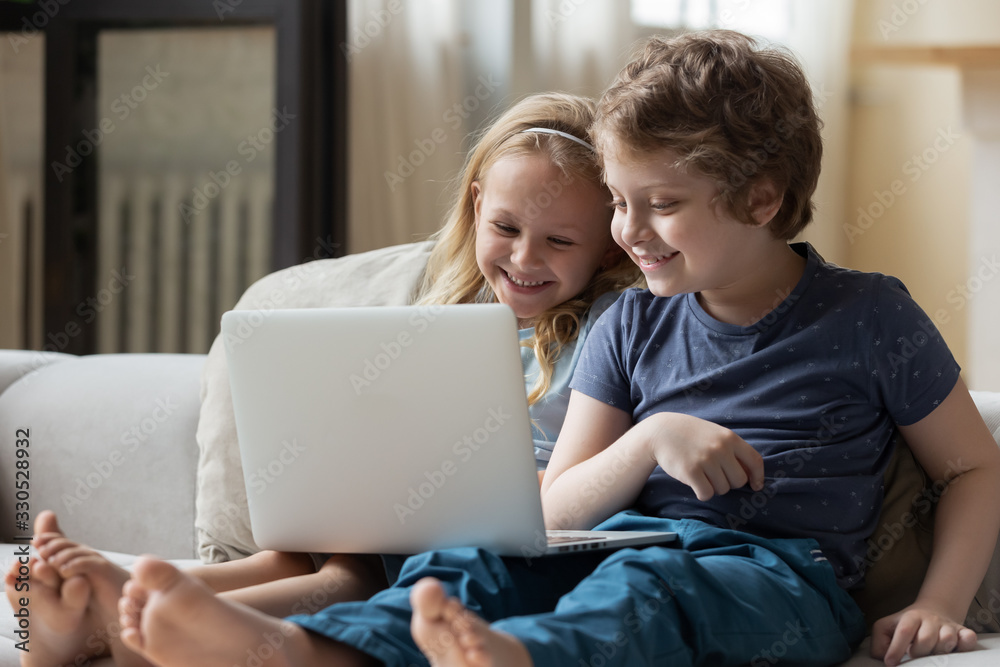 Funny smart small siblings sit on comfortable couch at home using laptop watching cartoon at home together, clever little kids brother and sister have fun enjoy movie or video on computer Funny smart small siblings sit on comfortable couch at home using laptop watching cartoon at home together, clever little kids brother and sister have fun enjoy movie or video on computer