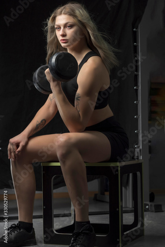 A girl with blond hair in a sports uniform is training with 10 kilogram dumbbells in the gym.