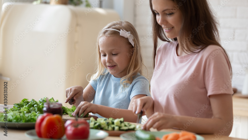 Close up of caring happy young mom enjoy leisure weekend time preparing salad chopping vegetables together with small daughter, loving mother teach little girl child cooking, healthy life concept