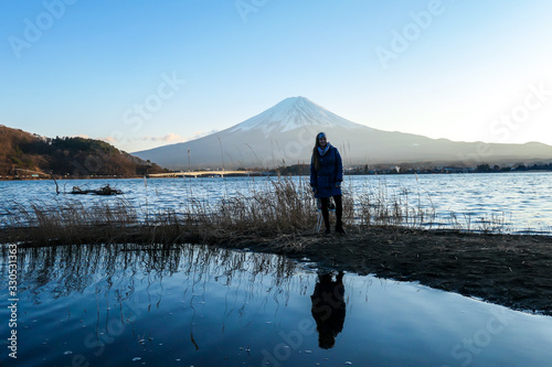 A woman walking at the side of Kawaguchiko Lake and watching Mt Fuji, Japan. Reflection of the woman in the water. Top of volcano covered with snow. Exploring new places. Serenity. Soft sunset colors