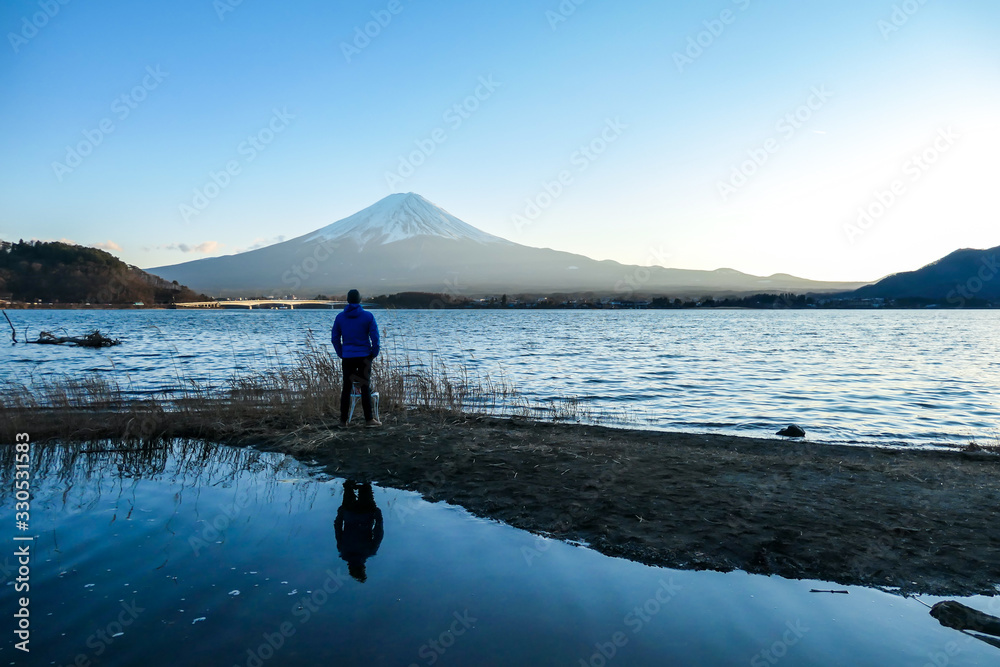 A man walking at the side of Kawaguchiko Lake and watching Mt Fuji, Japan. Reflection of the man in the water. Top of volcano covered with snow. Exploring new places. Soft sunset colors. Calmness A man walking at the side of Kawaguchiko Lake and watching Mt Fuji, Japan. Reflection of the man in the water. Top of volcano covered with snow. Exploring new places. Soft sunset colors. Calmness